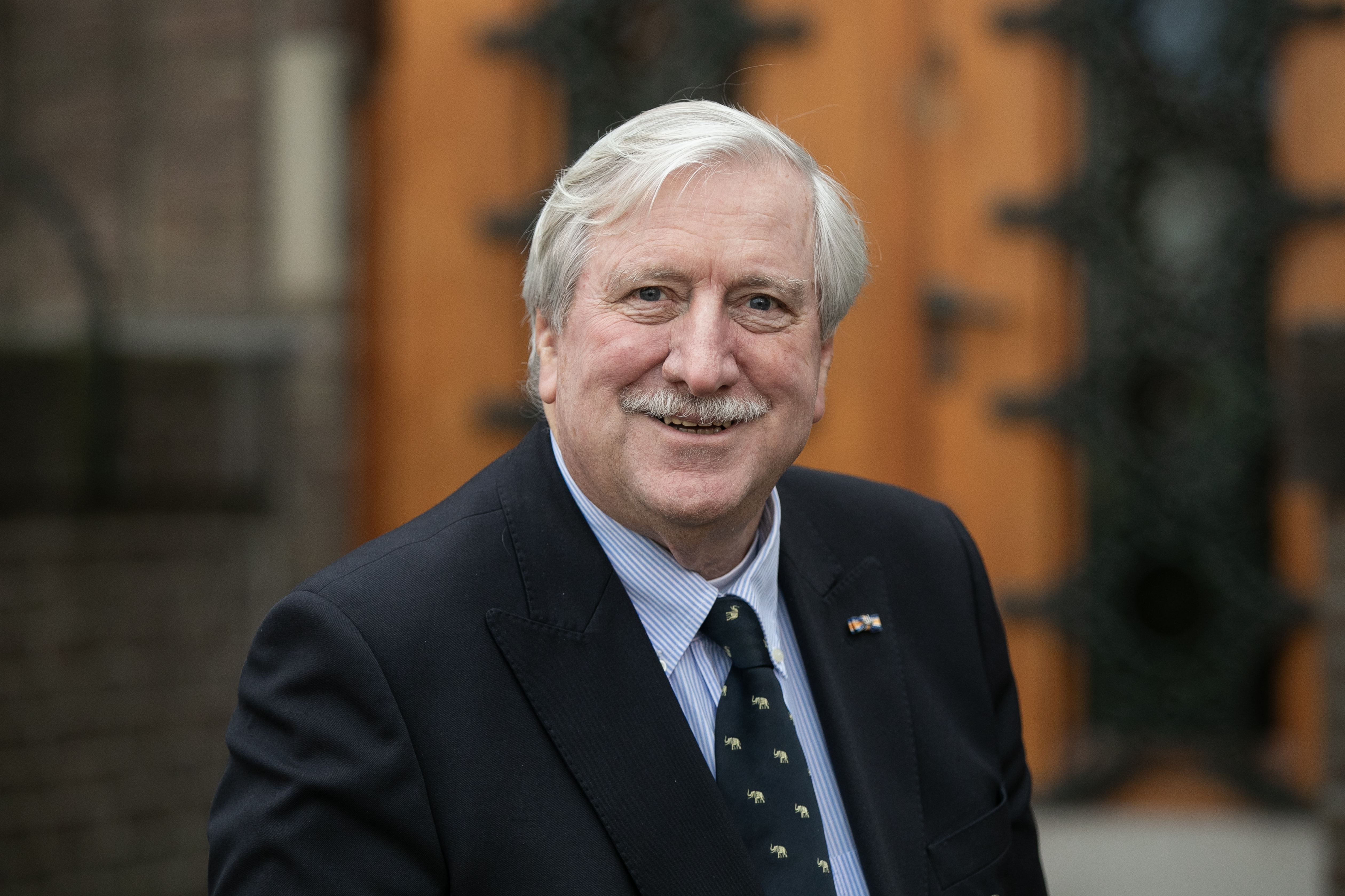 Een man met grijs haar en een snor, gekleed in een donker pak en een stropdas met patronen, glimlacht terwijl hij buiten staat met een houten deur op de achtergrond.