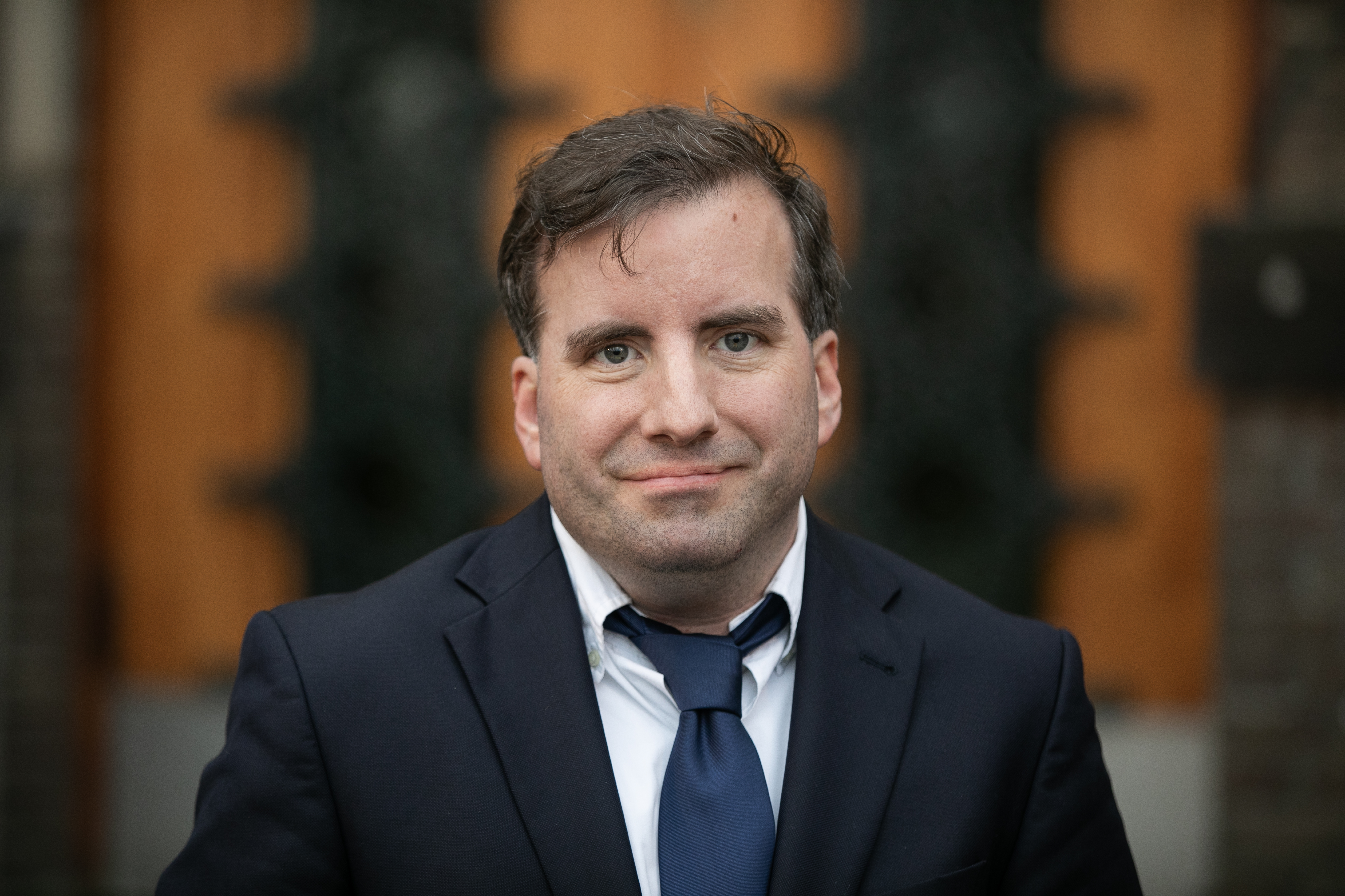 Portret van een man in een blazer met een das, staand voor een achtergrond van hout en een donker patroon.