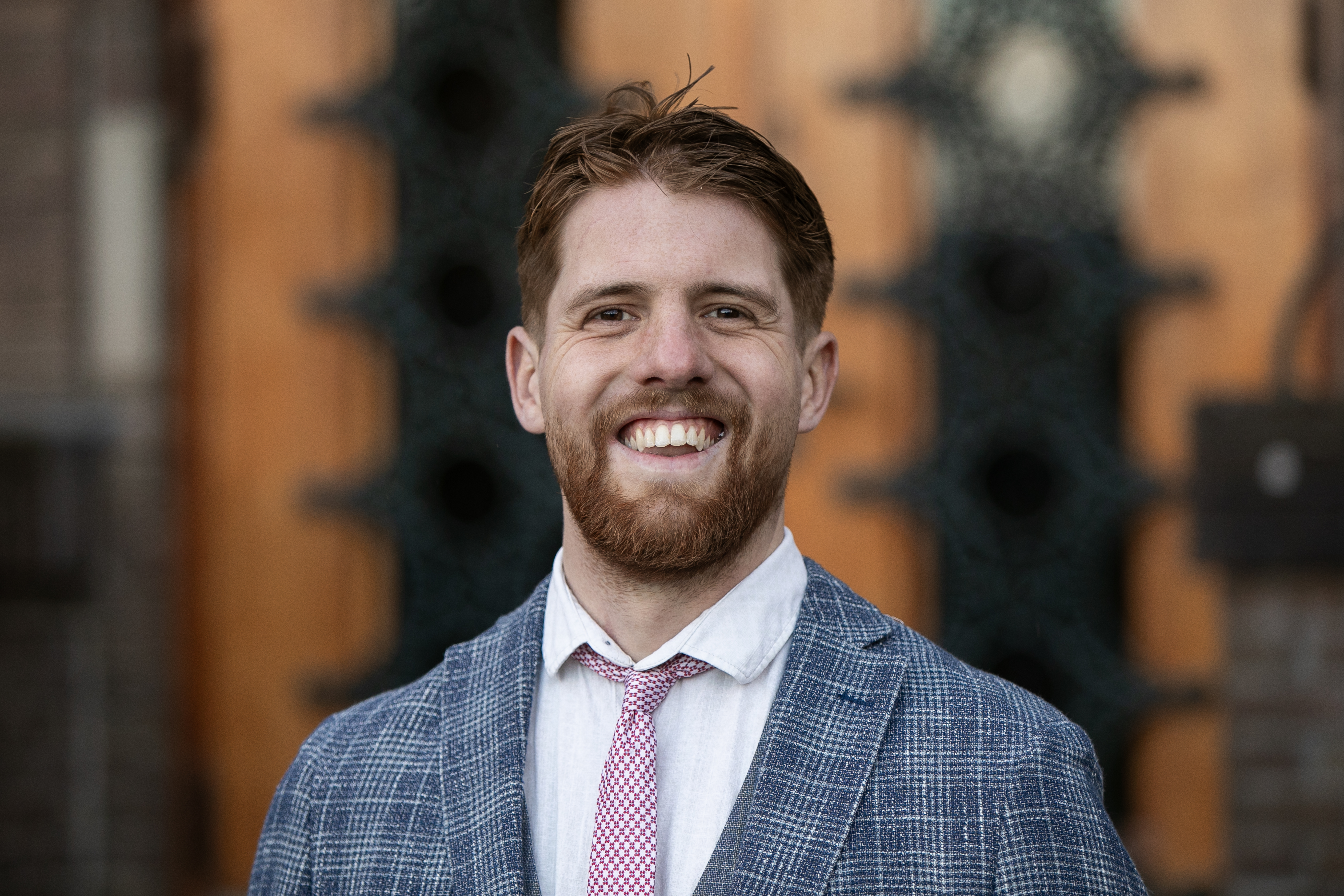 Een glimlachende man met een baard, gekleed in een blauw geruit jasje en een lichtgekleurde blouse, met een roze stropdas, staat voor een houten achtergrond.