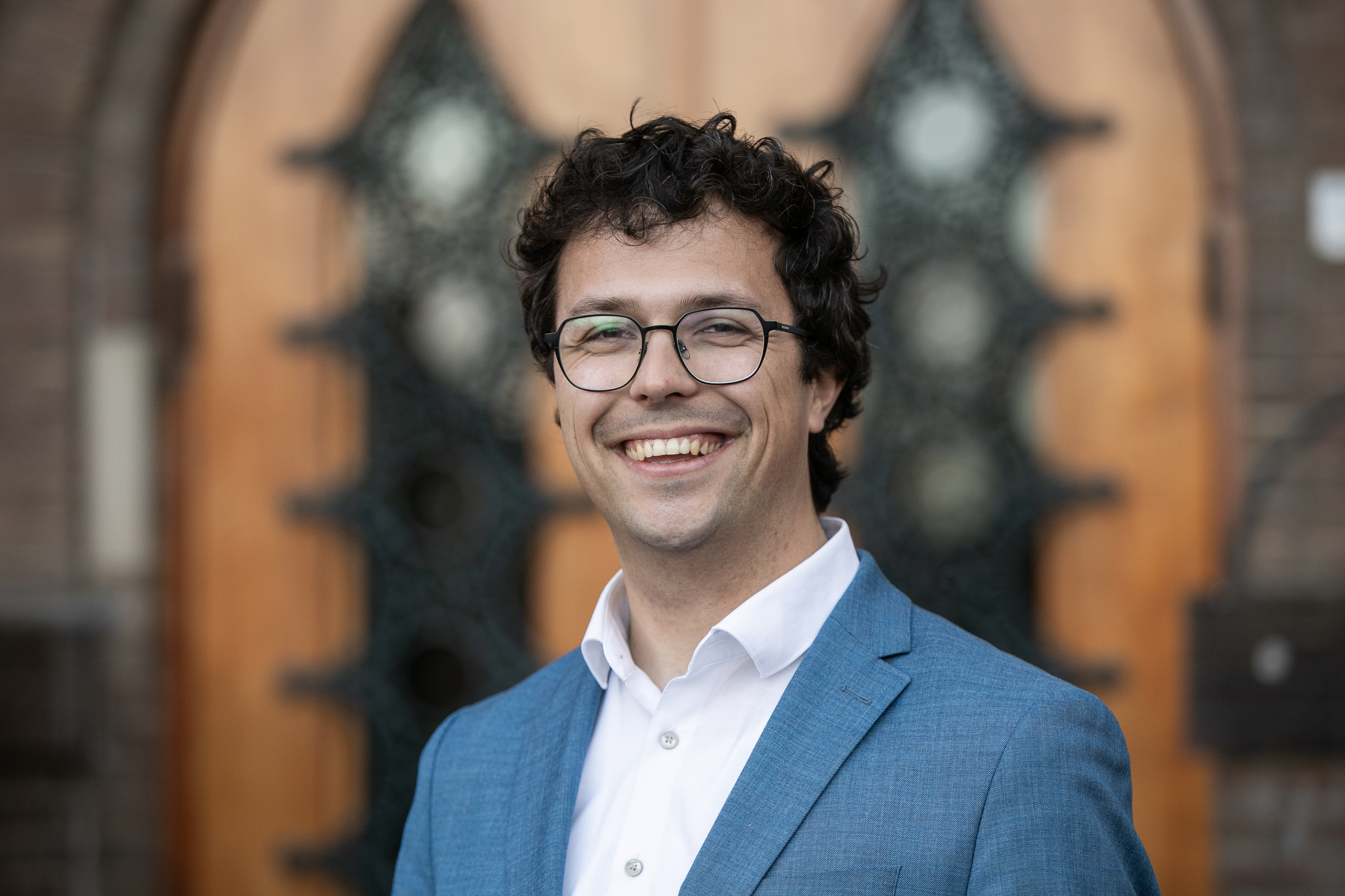 Man in een blauw jasje met een witte blouse, lachend voor een gebouw met een houten deur en decoratieve elementen op de achtergrond.