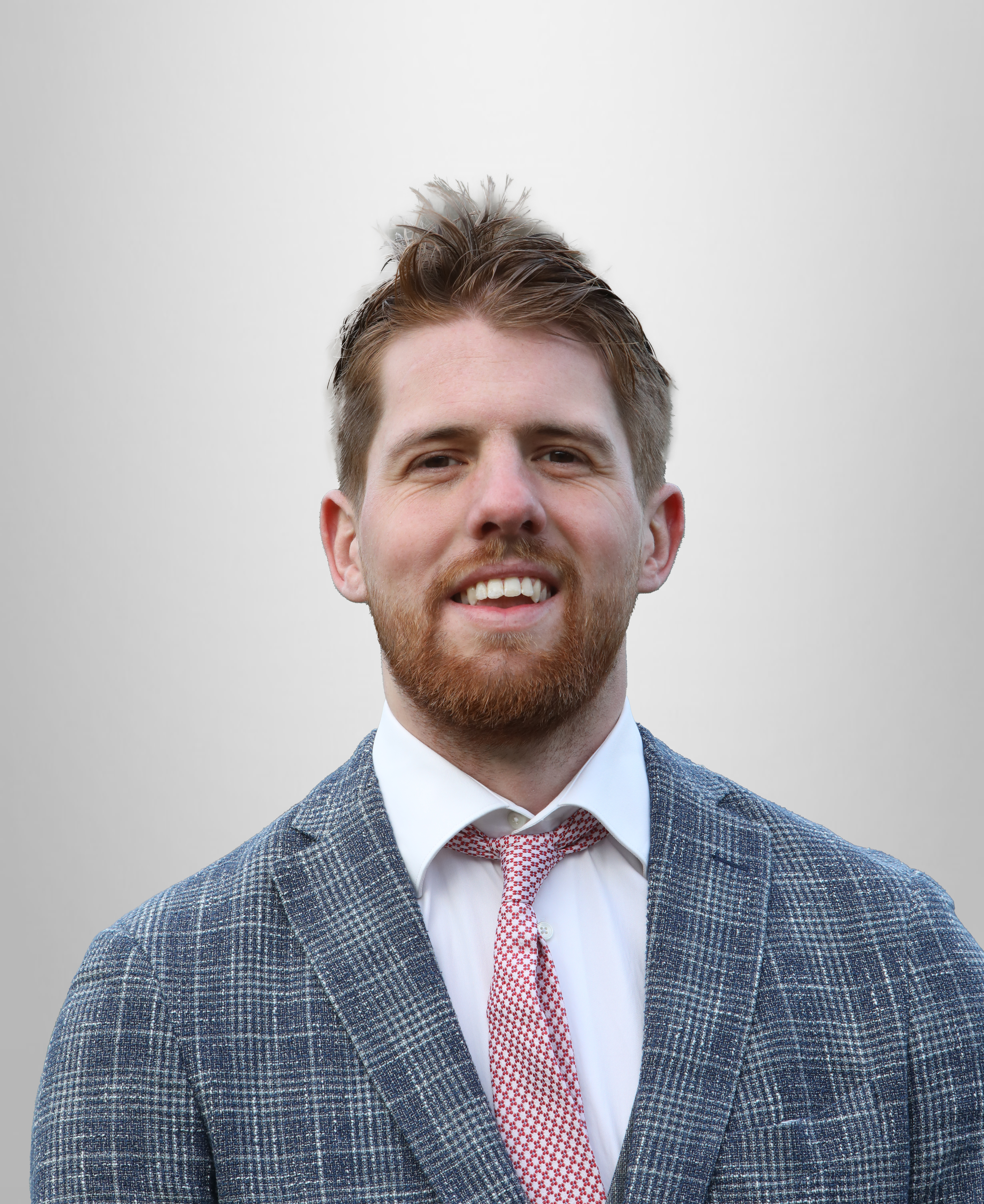 Een glimlachende man met een baard, gekleed in een grijs-blauw kostuum en een roze gestreepte das, staande voor een lichte achtergrond.